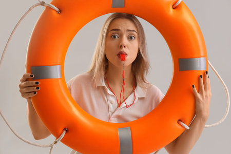 Female lifeguard with ring buoy whistling on light background