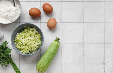 Bowls with grated zucchini, flour and eggs for preparing fritters on white tile backgroundの写真素材