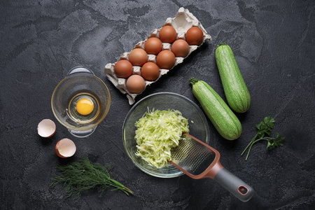 Bowl with grated zucchini and ingredients for preparing fritters on black backgroundの写真素材