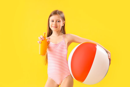 Little girl with bottle of sunscreen cream and inflatable ball on yellow backgroundの写真素材