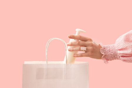 Hand of woman putting cosmetic product in shopping bag on pink backgroundの写真素材