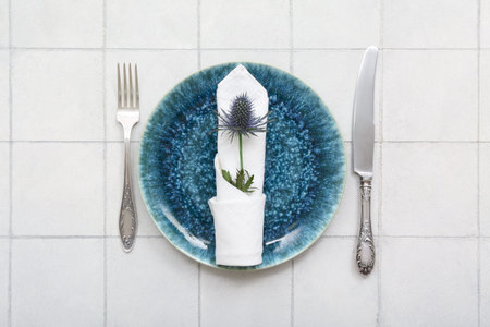 Beautiful table setting with clean plate, eryngium flower and cutlery on white tiled table