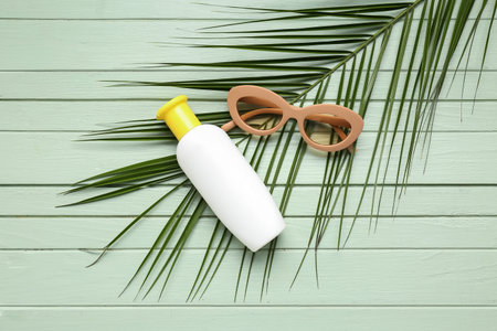 Sunglasses with bottle of sunscreen cream and palm leaf on green wooden background