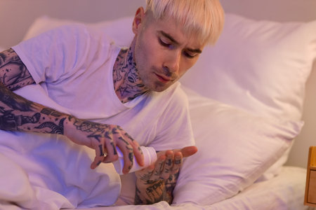 Young tattooed man taking sleeping pill in bed at nightの写真素材
