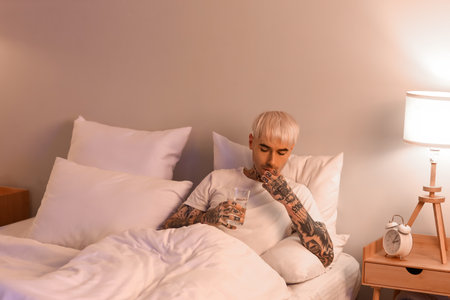 Young tattooed man with glass of water taking sleeping pill in bed at nightの写真素材