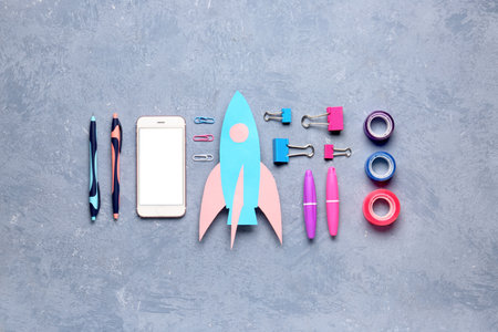 Blue paper rocket with mobile phone, pens and adhesive tape on gray grunge backgroundの写真素材