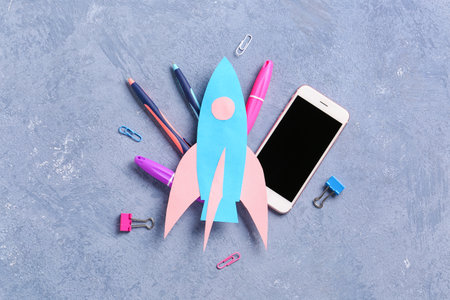 Blue paper rocket with mobile phone, pens and clips on gray grunge backgroundの写真素材