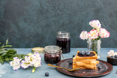 Plate with sweet blueberry jam toasts, jars and flowers on blue tableの写真素材