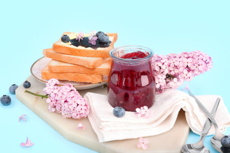 Board with jar of blueberry jam, toasts and flowers on blue background