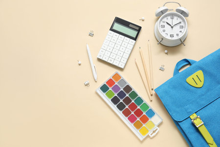 Blue school backpack with alarm clock, watercolors and calculator on beige background