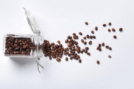 Glass jar with scattered coffee beans on white backgroundの写真素材