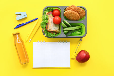 Stationery, blank notebook, drink and lunch box with tasty food on yellow backgroundの写真素材