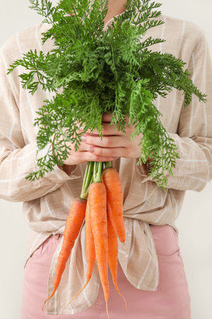 Woman holding bunch of fresh carrots with leaves on light background