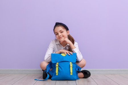 Little schoolgirl with backpack sitting near lilac wallの写真素材