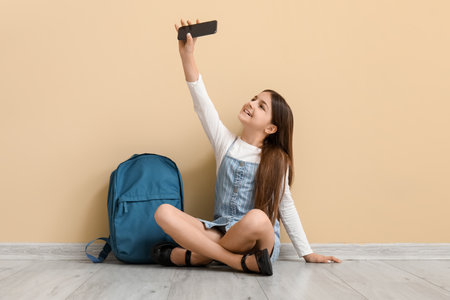 Little schoolgirl with backpack taking selfie near beige wallの写真素材