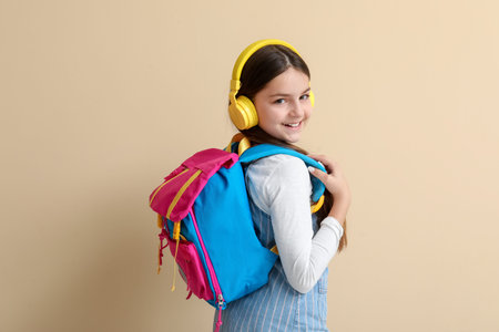 Little schoolgirl in headphones with backpack on beige background