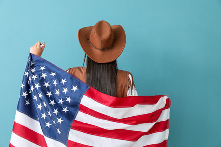 Beautiful young cowgirl holding USA flag on blue background, back viewの写真素材