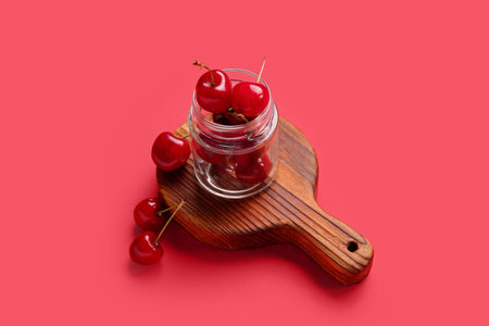 Wooden board with jar of sweet cherries on red background