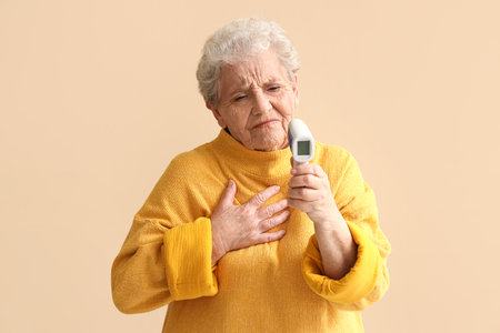 Ill senior woman with infrared thermometer on beige background