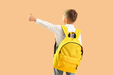 Little schoolboy with yellow backpack showing thumb-up gesture on orange background, back viewの写真素材