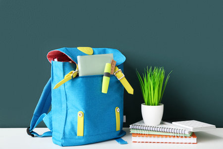 Blue school backpack with notebooks, calculator and houseplant on white wooden table near dark green...