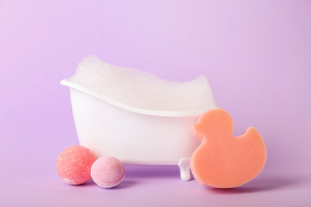 Small bathtub with foam, sponge and bath bombs on lilac backgroundの写真素材
