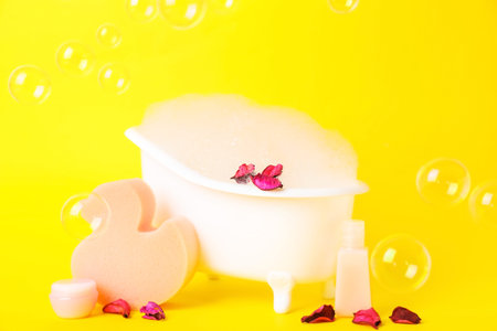 Small bathtub with foam, flower petals and bath supplies on yellow background
