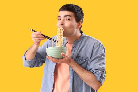 Young man eating Chinese noodles on yellow background