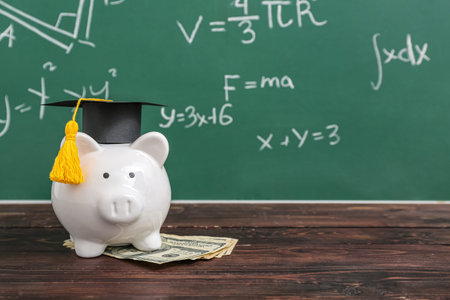 Piggy bank in graduation cap with dollar banknotes on brown wooden table against blackboard. student loan conceptの写真素材