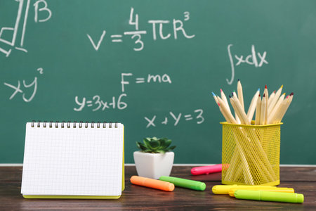 Blank notebook and stationery on wooden table against green chalkboard with formulasの写真素材