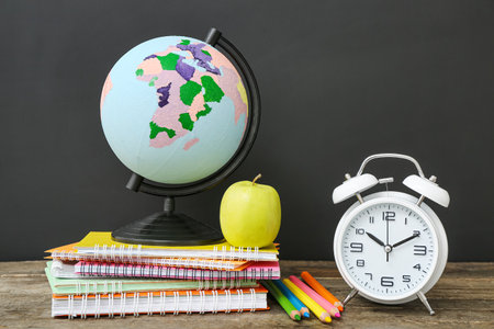 Different stationery with globe and alarm clock on wooden table against black chalkboardの写真素材
