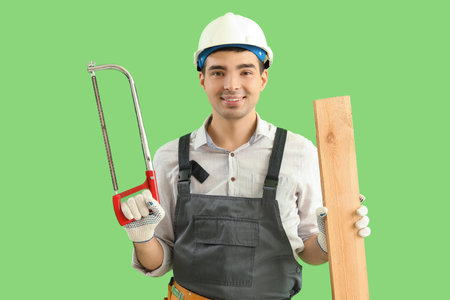 Young carpenter with hacksaw and wooden plank on green background