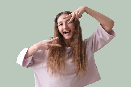 Beautiful young woman in pink sweatshirt showing victory gesture on green background