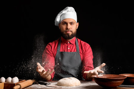 Male baker sprinkling dough with flour at table on dark backgroundの写真素材