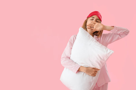 Young woman in pajamas with pillow yawning on pink backgroundの写真素材