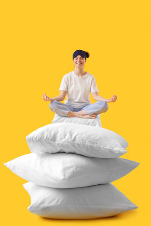 Young woman with sleeping mask and pillows meditating on yellow background
