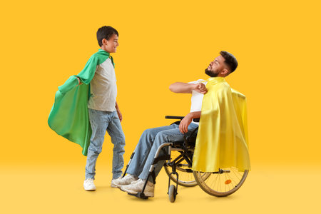 Little boy and his father in wheelchair wearing superhero capes on yellow backgroundの写真素材