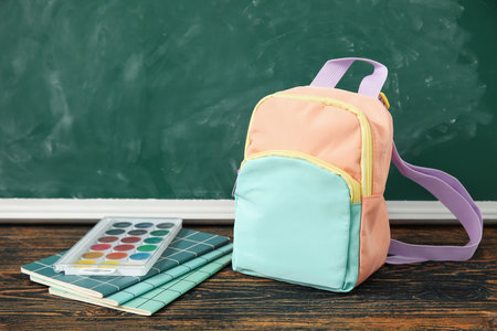 Colorful school backpack with notebooks and watercolors on wooden table near green chalkboardの写真素材