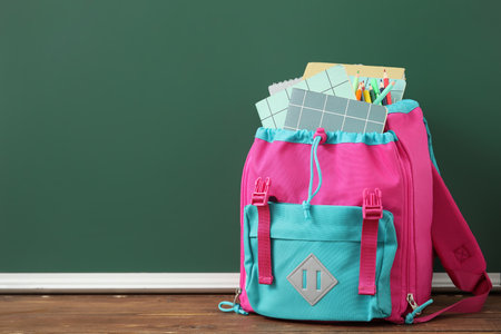 Colorful school backpack with notebooks and pencils on wooden table near green chalkboardの写真素材