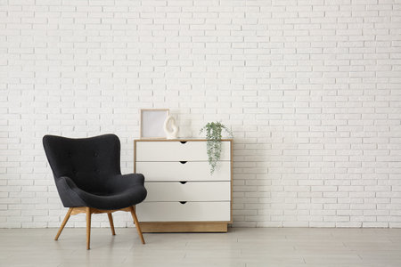 Stylish black armchair and chest of drawers near white brick wallの写真素材