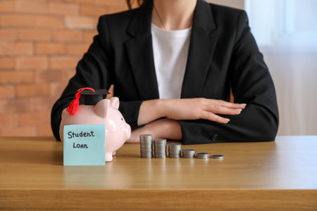 Woman in business suit sitting at table with piggy bank, stacks of coins and text STUDENT LOAN on sticky noteの写真素材