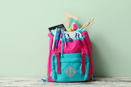 Colorful school backpack with different stationery on wooden table near pale green wall