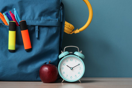 Colorful school backpack with different stationery and alarm clock on wooden table near blue wall