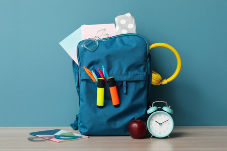 Colorful school backpack with different stationery and alarm clock on wooden table near blue wall