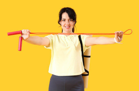 Sporty young woman with jumping rope and yoga mat on yellow background