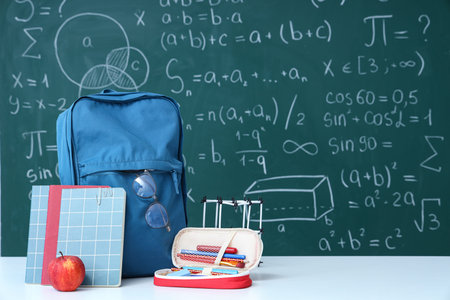 Blue school backpack with fresh apple and stationery on white table near green chalkboard