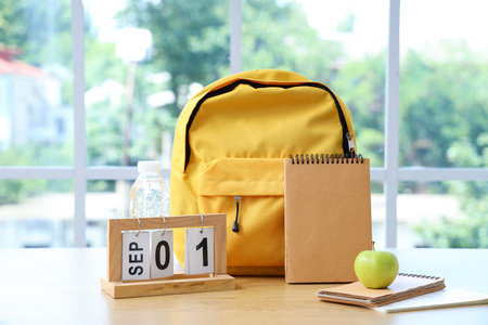 Yellow school backpack with stationery and calendar on brown wooden table near window in classroom