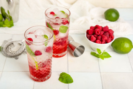 Glasses of fresh raspberry mojito and bowl with berries on white tile tableの写真素材