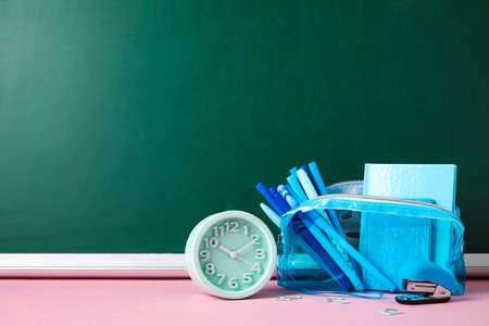 Pencil case with different school stationery and alarm clock on pink table near blackboardの写真素材