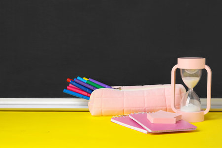 Pencil case with different school stationery and hourglass on yellow table near blackboard